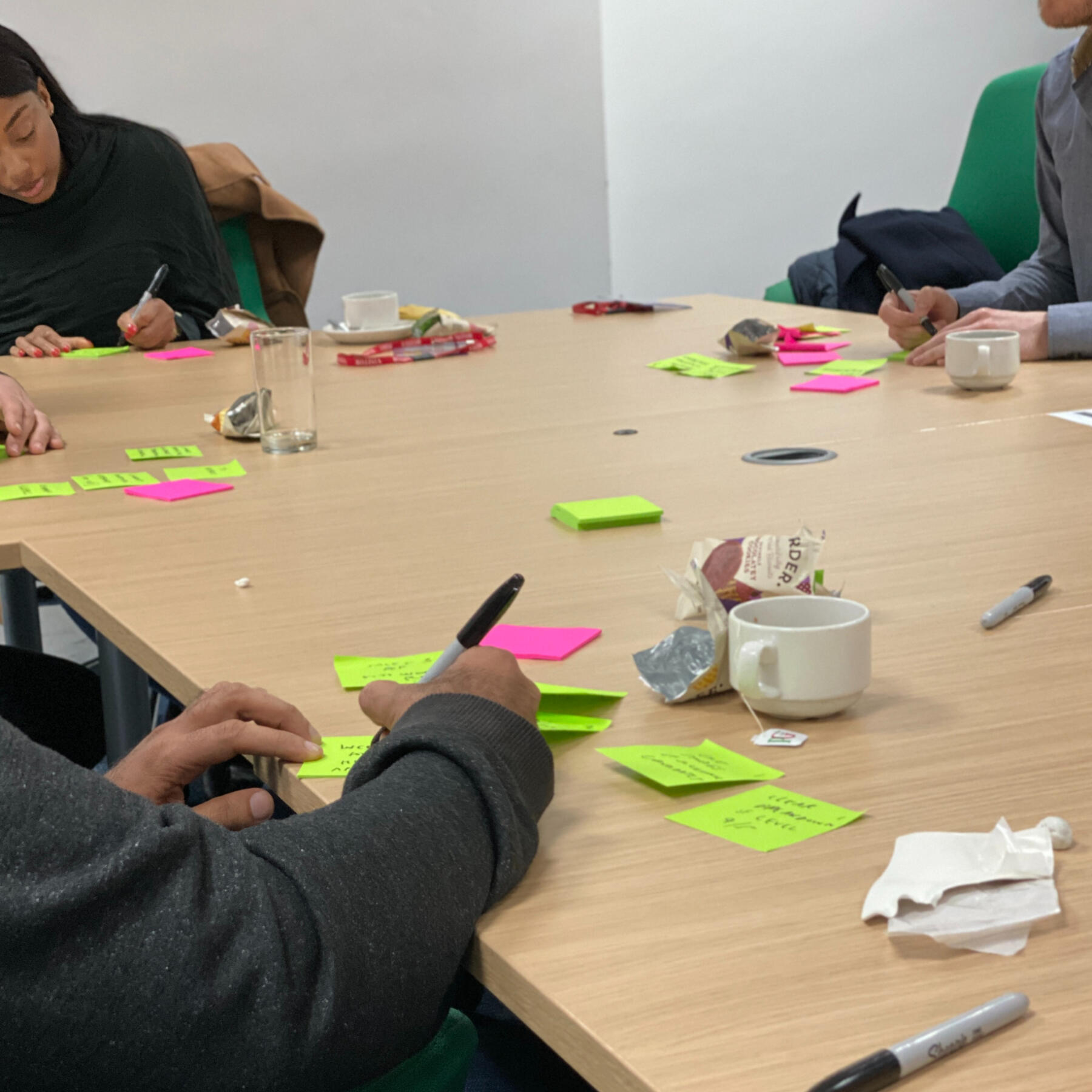 Participants seated around a table, engaged in a workshop activity. Bright neon sticky notes, markers, and coffee cups are spread across the table as people write down ideas.