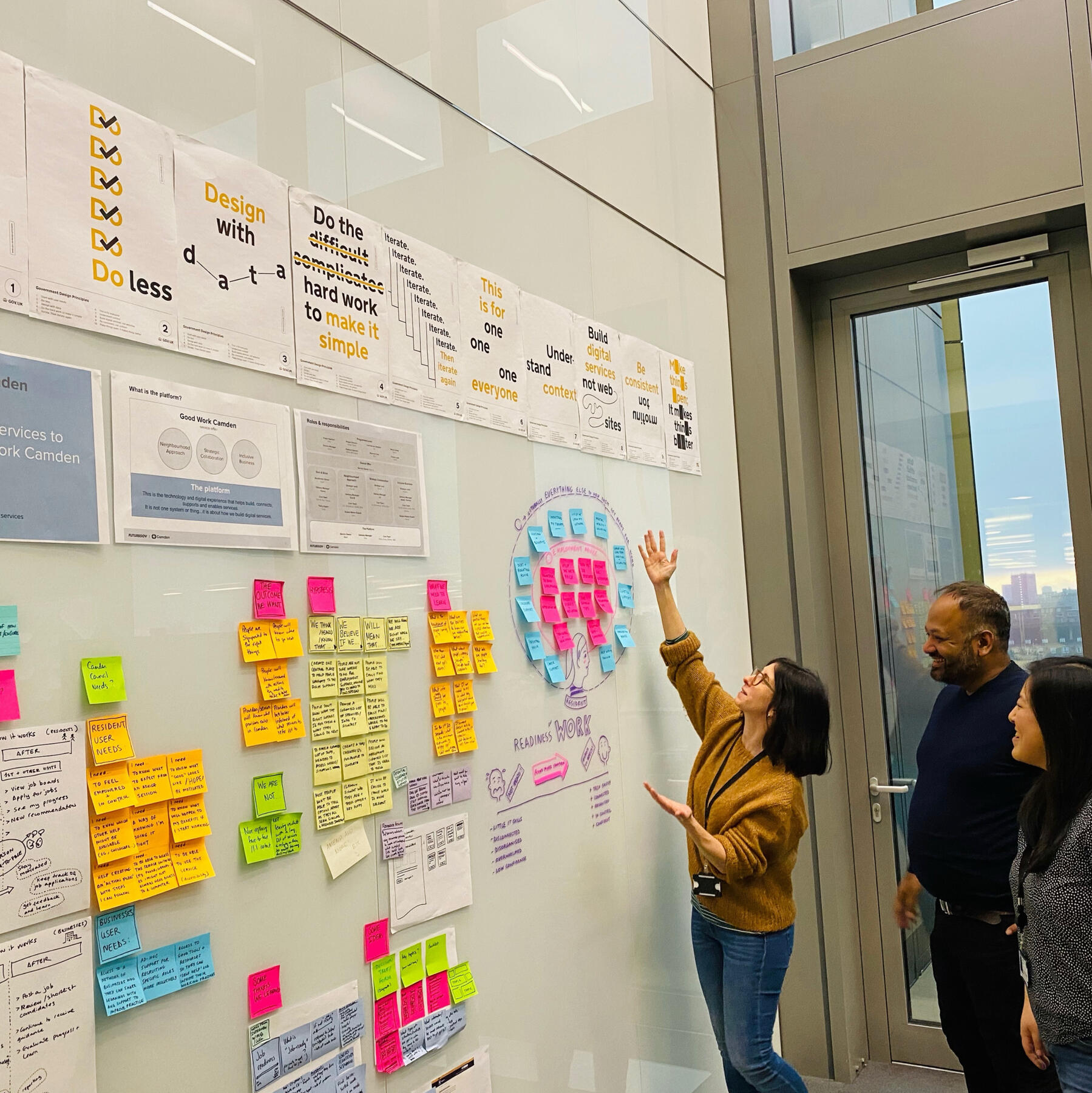 Mar standing in front of a wall covered with sticky notes, sketches, and design principles, explaining ideas to two colleagues during a workshop. The wall is filled with colorful notes and diagrams related to the project at hand.